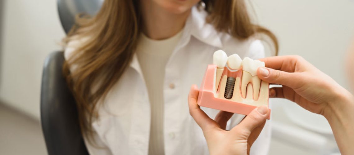 First-person-shot-female-doctor-talking-to-a-young-woman-patient.-Discussion-of-the-treatment-plan.-The-dentist-shows-a-model-of-a-dental-implant