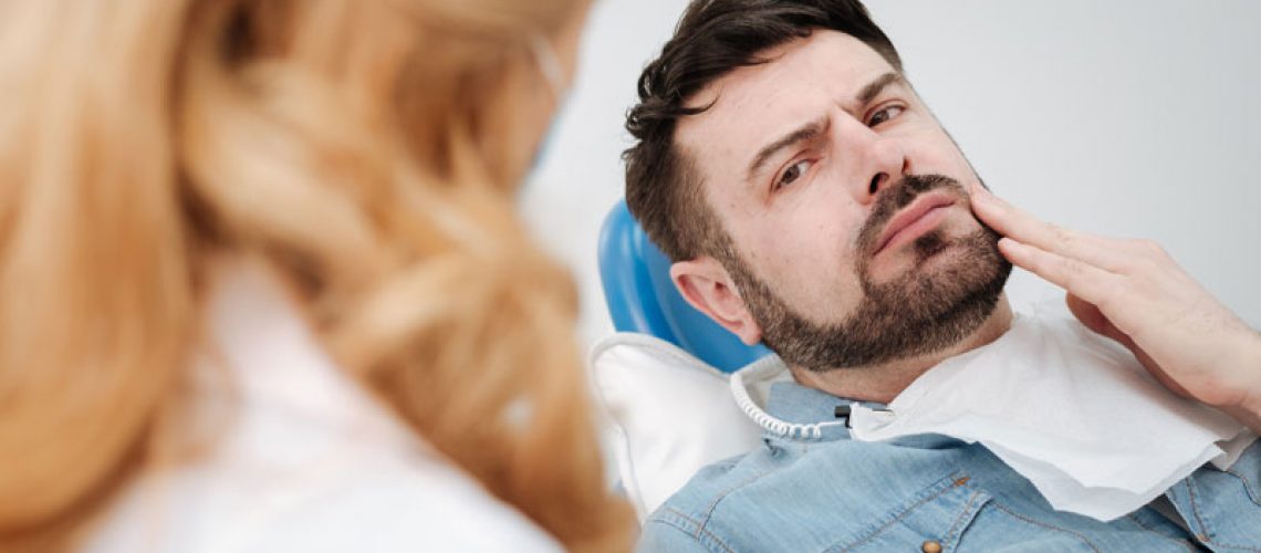 a dental patient touching the side of his face in pain as an oral surgeon explains what could happen to him if he does not get his wisdom teeth removed.