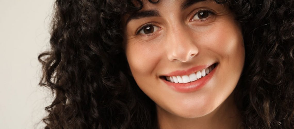Confident young woman with curly hair smiling brightly, showing straight white teeth against a soft light grey background.